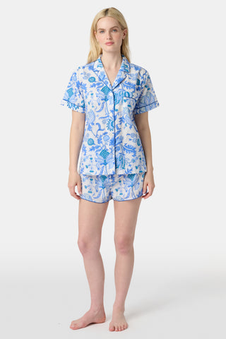 A woman stands in front of lush greenery, hands in her pockets and looking relaxed, wearing a straw hat and the Amanda Kurta—a blue-and-white floral piece made from 100% cotton.