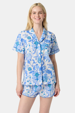 A woman stands in front of lush greenery, hands in her pockets and looking relaxed, wearing a straw hat and the Amanda Kurta—a blue-and-white floral piece made from 100% cotton.