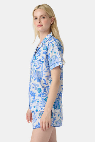 A woman stands in front of lush greenery, hands in her pockets and looking relaxed, wearing a straw hat and the Amanda Kurta—a blue-and-white floral piece made from 100% cotton.