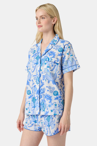 A woman stands in front of lush greenery, hands in her pockets and looking relaxed, wearing a straw hat and the Amanda Kurta—a blue-and-white floral piece made from 100% cotton.