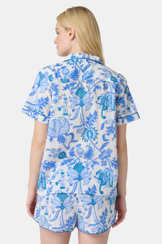A woman stands in front of lush greenery, hands in her pockets and looking relaxed, wearing a straw hat and the Amanda Kurta—a blue-and-white floral piece made from 100% cotton.