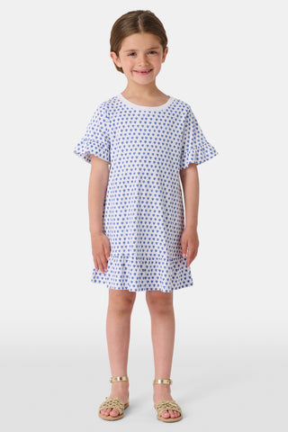 A young girl wearing a Roller Rabbit Girls Hearts Esme Dress stands between two light blue bikes, holding an adult’s hand. She has a large white bow in her hair and gazes thoughtfully to the side.