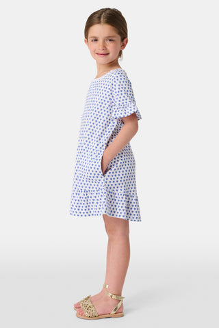 A young girl wearing a Roller Rabbit Girls Hearts Esme Dress stands between two light blue bikes, holding an adult’s hand. She has a large white bow in her hair and gazes thoughtfully to the side.