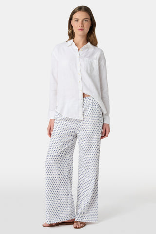 A person stands facing forward, wearing Roller Rabbit’s Hearts Kaylee Pant and a white cotton button-up shirt with brown flat sandals against a plain light background.