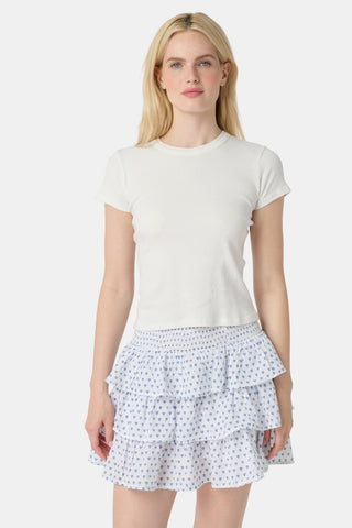 A woman with long blonde hair wears a fitted white tee and the Roller Rabbit Hearts Siggy Skirt, a white ruffle mini skirt with a blue hearts print, standing against a plain white background.