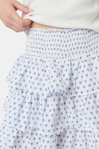 A woman with long blonde hair wears a fitted white tee and the Roller Rabbit Hearts Siggy Skirt, a white ruffle mini skirt with a blue hearts print, standing against a plain white background.