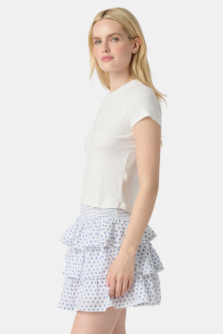 A woman with long blonde hair wears a fitted white tee and the Roller Rabbit Hearts Siggy Skirt, a white ruffle mini skirt with a blue hearts print, standing against a plain white background.