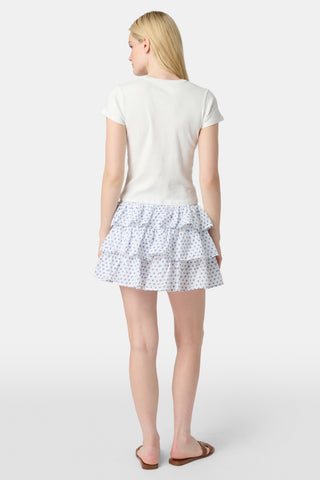 A woman with long blonde hair wears a fitted white tee and the Roller Rabbit Hearts Siggy Skirt, a white ruffle mini skirt with a blue hearts print, standing against a plain white background.