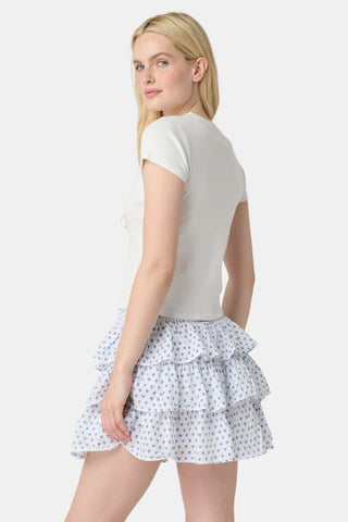 A woman with long blonde hair wears a fitted white tee and the Roller Rabbit Hearts Siggy Skirt, a white ruffle mini skirt with a blue hearts print, standing against a plain white background.