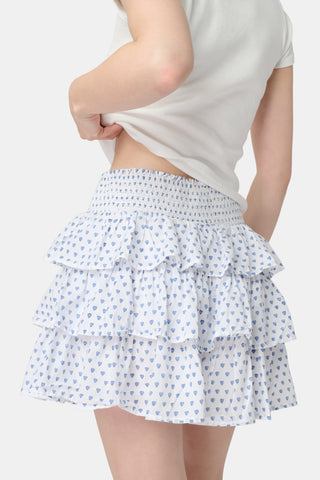 A woman with long blonde hair wears a fitted white tee and the Roller Rabbit Hearts Siggy Skirt, a white ruffle mini skirt with a blue hearts print, standing against a plain white background.