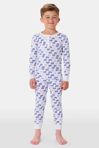 A girl stands barefoot, smiling, in Roller Rabbit Kids Moby Pajamas—a matching white long-sleeve set with blue smiling face pattern, buttoned neckline, and fitted style. The background is plain light gray.