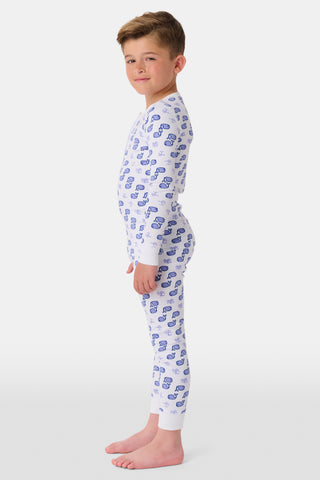 A girl stands barefoot, smiling, in Roller Rabbit Kids Moby Pajamas—a matching white long-sleeve set with blue smiling face pattern, buttoned neckline, and fitted style. The background is plain light gray.
