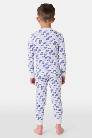 A girl stands barefoot, smiling, in Roller Rabbit Kids Moby Pajamas—a matching white long-sleeve set with blue smiling face pattern, buttoned neckline, and fitted style. The background is plain light gray.