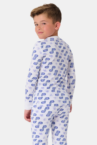 A girl stands barefoot, smiling, in Roller Rabbit Kids Moby Pajamas—a matching white long-sleeve set with blue smiling face pattern, buttoned neckline, and fitted style. The background is plain light gray.