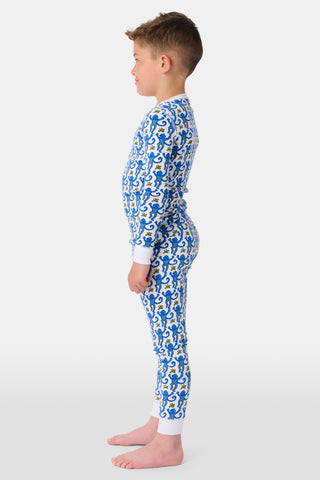 A young girl with curly hair stands barefoot in Kids Monkey Pajamas—a matching long-sleeve top and pants set made from soft 100% cotton, featuring playful monkey prints, against a plain white background.