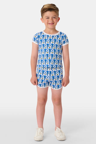 A young child in the Kids Monkey Playa Play Set—blue patterned cotton pajamas with playful Roller Rabbit monkeys—walks hand-in-hand with an adult in pink pajamas outside near green grass, while another brown-haired child sits nearby.