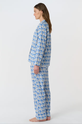 A woman smiles in the Monkey Loungewear long sleeve pajama set by Roller Rabbit, featuring a blue and white floral pattern. She stands barefoot with hands on hips against a plain white background.
