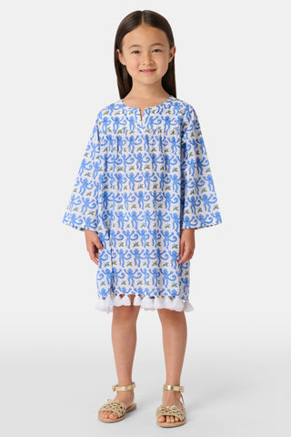A young girl with long dark hair wears a Roller Rabbit Kids Monkey Serafina Tunic, a blue-and-white cotton voile dress with long sleeves and white fringe at the hem, standing against a plain white background.