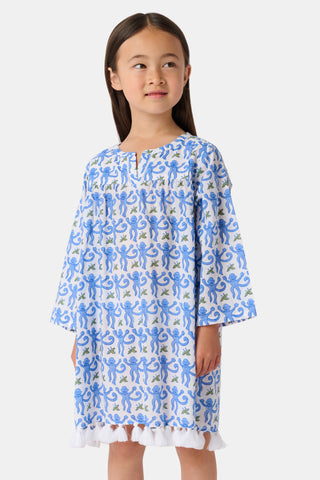 A young girl with long dark hair wears a Roller Rabbit Kids Monkey Serafina Tunic, a blue-and-white cotton voile dress with long sleeves and white fringe at the hem, standing against a plain white background.