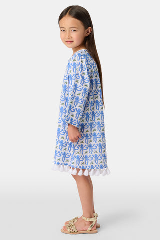 A young girl with long dark hair wears a Roller Rabbit Kids Monkey Serafina Tunic, a blue-and-white cotton voile dress with long sleeves and white fringe at the hem, standing against a plain white background.