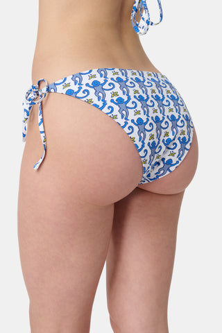 A person wears Roller Rabbit's Monkey/Hearts Serena Bikini Bottom in blue and white with monkey print and side ties. Only the torso and upper legs are visible against a plain background. This reversible bikini bottom provides UPF 50+ sun protection.