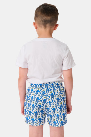 A child stands barefoot in a white t-shirt and Roller Rabbit Boys Monkey Swim Trunks, featuring a playful blue monkey print, with hands in pockets against a plain white background.