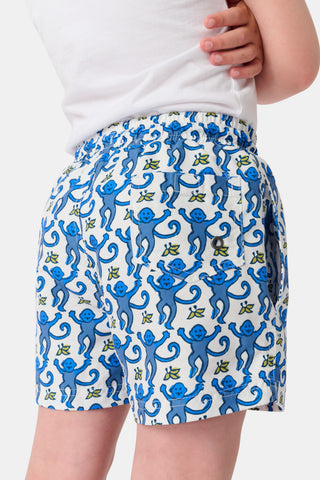 A child stands barefoot in a white t-shirt and Roller Rabbit Boys Monkey Swim Trunks, featuring a playful blue monkey print, with hands in pockets against a plain white background.