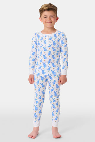 A young child with curly hair stands barefoot in white Kids Romy Rabbit Pajamas by Roller Rabbit, featuring a blue bunny print, looking at the camera and touching their mouth with one finger.