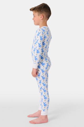 A young child with curly hair stands barefoot in white Kids Romy Rabbit Pajamas by Roller Rabbit, featuring a blue bunny print, looking at the camera and touching their mouth with one finger.