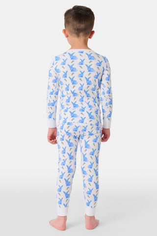 A young child with curly hair stands barefoot in white Kids Romy Rabbit Pajamas by Roller Rabbit, featuring a blue bunny print, looking at the camera and touching their mouth with one finger.