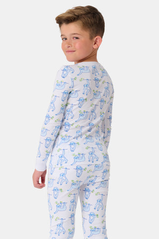 A young boy stands barefoot, smiling and facing forward in Roller Rabbit's Kids Sleepy Sloth Pajamas—a white, long-sleeve cotton PJ set with playful blue and green sloth prints—against a plain white background.