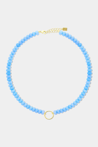 The Beaded Charm Necklace by Roller Rabbit features round, evenly spaced blue beads with a gold clasp, accented by a small gold ring pendant at the center. The necklace is shown against a plain white background.