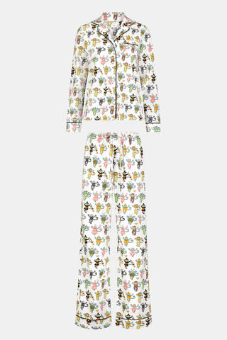 A woman stands barefoot against a plain white background, wearing the Roller Rabbit Boo Crew Long Sleeve Polo Pajamas, which feature a colorful cartoon bee and flower pattern on white Pima cotton with long sleeves and button-up styling.