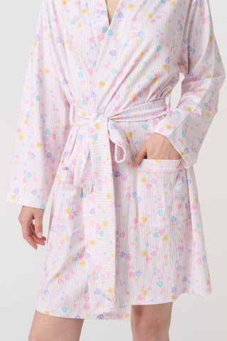 A woman with blonde hair models the Roller Rabbit Candy Heart Stripe Blake Robe—a light pink, knee-length Pima cotton robe with candy heart stripes and a waist tie—against a plain white background.