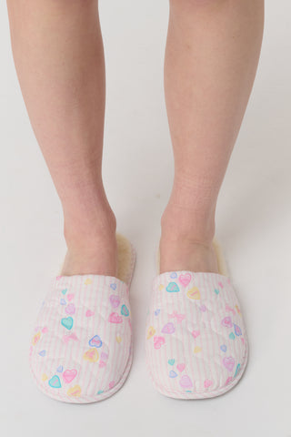 A person stands on a white surface wearing Roller Rabbit Candy Heart Stripe Quilted Slippers decorated with colorful heart patterns.