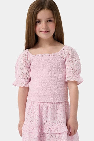 A young girl with long brown hair is wearing the Roller Rabbit Kids Clover Eyelet Elsie Top, a light pink, short-sleeved, smocked kids top, paired with a matching heart-patterned eyelet skirt. She smiles against a plain light background.