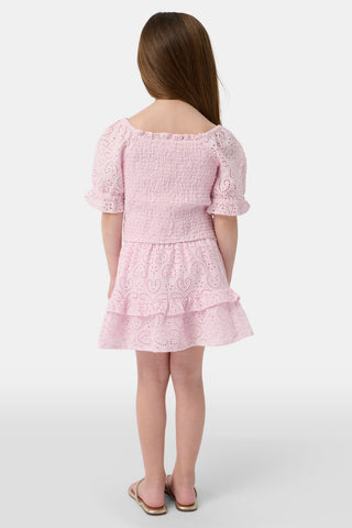 A young girl with long brown hair is wearing the Roller Rabbit Kids Clover Eyelet Elsie Top, a light pink, short-sleeved, smocked kids top, paired with a matching heart-patterned eyelet skirt. She smiles against a plain light background.