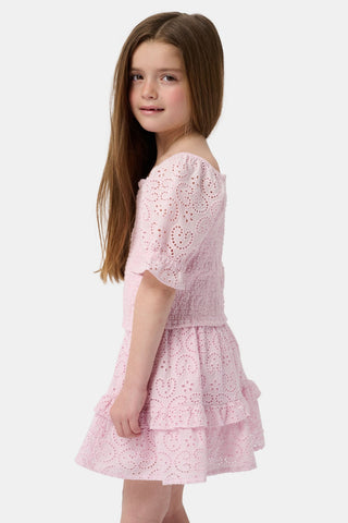 A young girl with long brown hair is wearing the Roller Rabbit Kids Clover Eyelet Elsie Top, a light pink, short-sleeved, smocked kids top, paired with a matching heart-patterned eyelet skirt. She smiles against a plain light background.