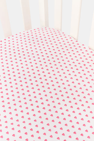 A close-up of the Roller Rabbit Hearts Crib Sheet in white, featuring a pattern of small pink hearts. In the background, part of the vertical back slats of a white chair is visible.
