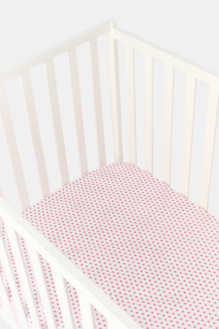 A close-up of the Roller Rabbit Hearts Crib Sheet in white, featuring a pattern of small pink hearts. In the background, part of the vertical back slats of a white chair is visible.