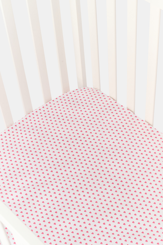 A close-up of the Roller Rabbit Hearts Crib Sheet in white, featuring a pattern of small pink hearts. In the background, part of the vertical back slats of a white chair is visible.