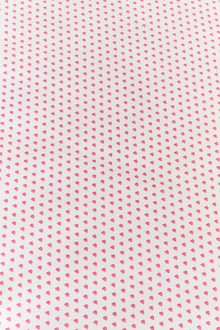 A close-up of the Roller Rabbit Hearts Crib Sheet in white, featuring a pattern of small pink hearts. In the background, part of the vertical back slats of a white chair is visible.
