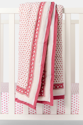 A white crib features pink heart-patterned sheets, with the Roller Rabbit Hearts Baby Quilt—a soft cotton quilt with a hearts print and decorative border—draped over the rail, adding charm to the bedding.