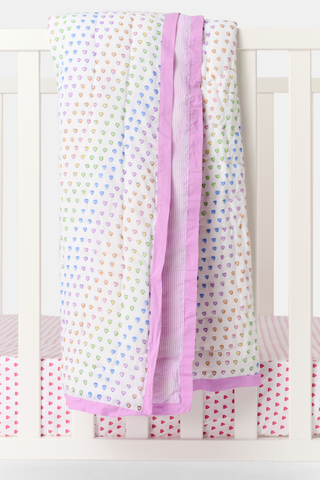 The Roller Rabbit Disco Hearts Baby Quilt, white cotton with colorful hearts and a light purple border, is draped over a white crib rail; the crib mattress has a pink and white striped sheet with small red hearts.