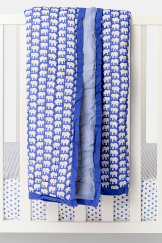 The Hathi Baby Quilt is a cotton quilt with a folded design, featuring a blue background with repeating white elephants, a contrasting striped section, and a solid blue border.