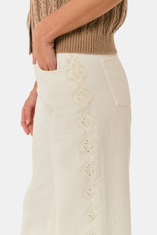Wearing Roller Rabbit’s Elsa Eyelet Danielle Jean—a high-waisted, wide-leg style in ecru with delicate scalloped lace trim along the sides—paired with a white top and black Mary Jane shoes, the individual stands against a plain white background.