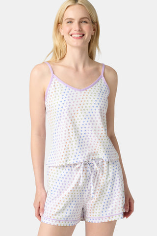 A woman with long blonde hair wears the Roller Rabbit Disco Hearts Mischa Set, a Pima cotton loungewear featuring a heart print on white. The set includes a sleeveless top with lavender straps and drawstring shorts. She smiles against a plain backdrop.