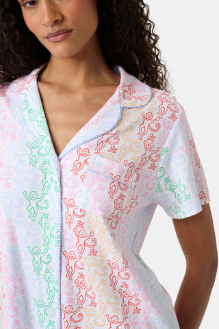 A woman with curly hair, standing and smiling slightly, wears Roller Rabbit’s Disco Monkey Wyatt Polo Pajamas: a short-sleeve button-up top and matching ruffled shorts featuring a pastel multicolor abstract pattern on white.