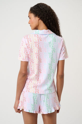 A woman with curly hair, standing and smiling slightly, wears Roller Rabbit’s Disco Monkey Wyatt Polo Pajamas: a short-sleeve button-up top and matching ruffled shorts featuring a pastel multicolor abstract pattern on white.