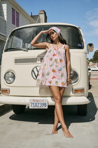 A woman in the Roller Rabbit Fruits D'Ete Keaton Dress with a matching headscarf stands barefoot by a vintage white Volkswagen van, shielding her eyes from the sun. The sunny coastal backdrop highlights the dress's playful print.